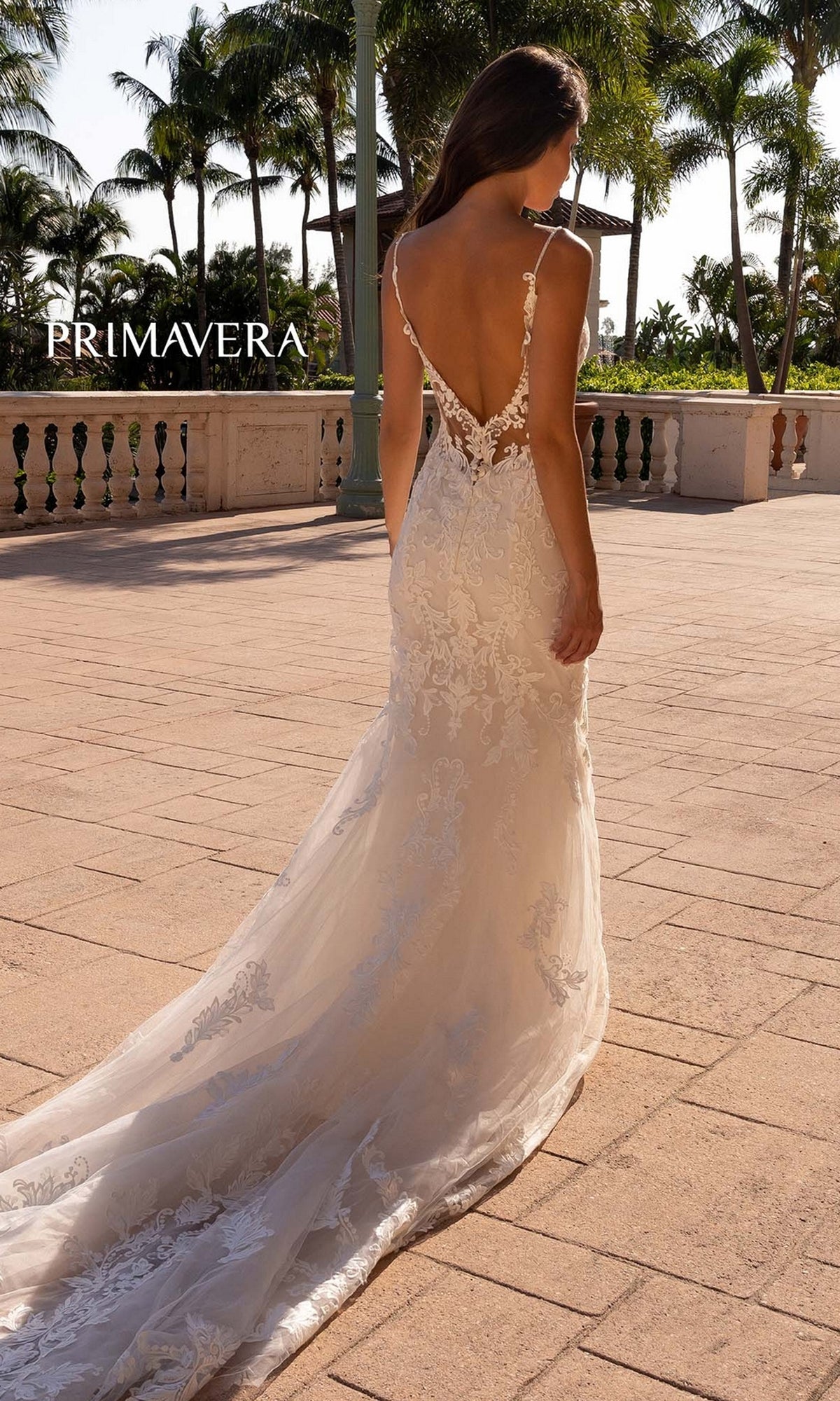 A woman stands outdoors on a sunlit terrace with palm trees, wearing the Primavera Long Wedding Dress 11137—an ivory, fitted gown featuring lace embroidery and a long train.